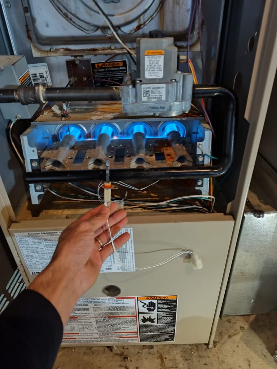 Hand holding a sensor wire in front of an active furnace burner assembly with blue flames