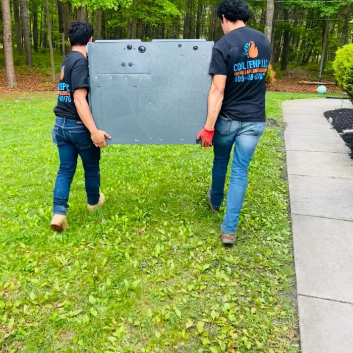 Two people carrying a large gray object across a grassy lawn with trees in the background.