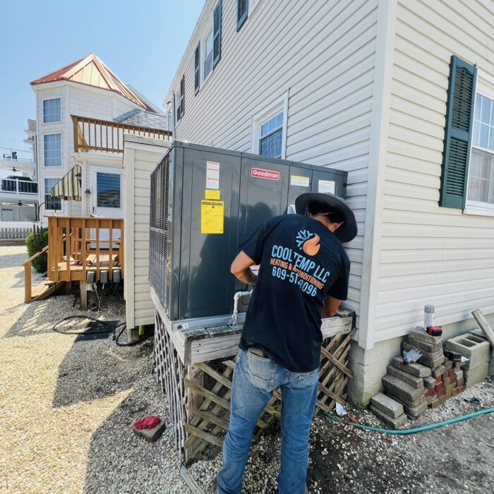 Technician in a black t-shirt works on an outdoor HVAC unit beside a beige house on a sunny day.