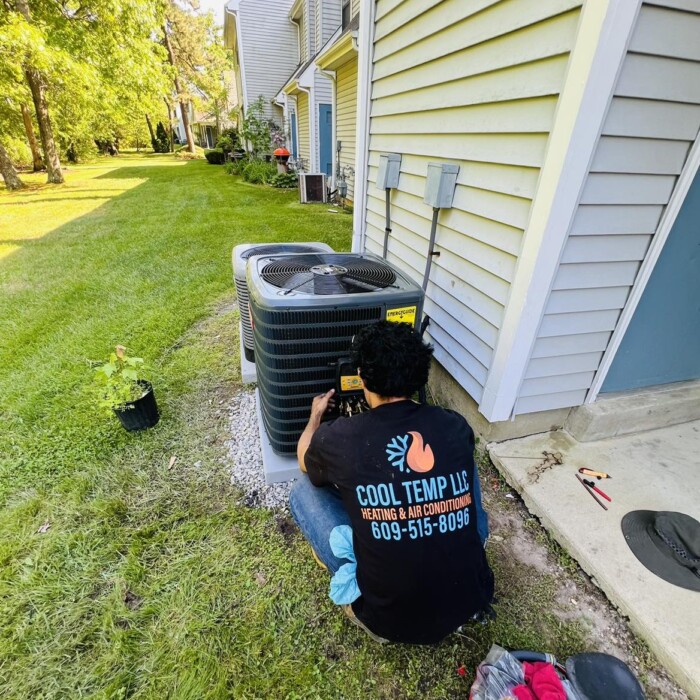 Technician repairs an outdoor air conditioning unit beside a house on a grassy lawn. Toolbox and equipment are nearby.